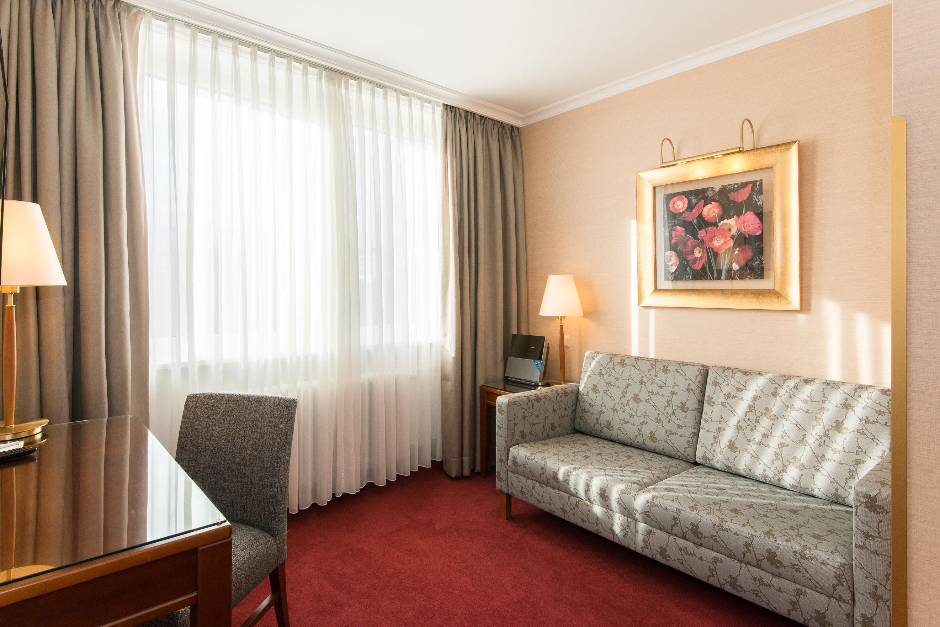 Ein Hotelzimmer mit Sofa, Stuhl, Schreibtisch, Lampe, Laptop und einem Gemälde an der Wand.