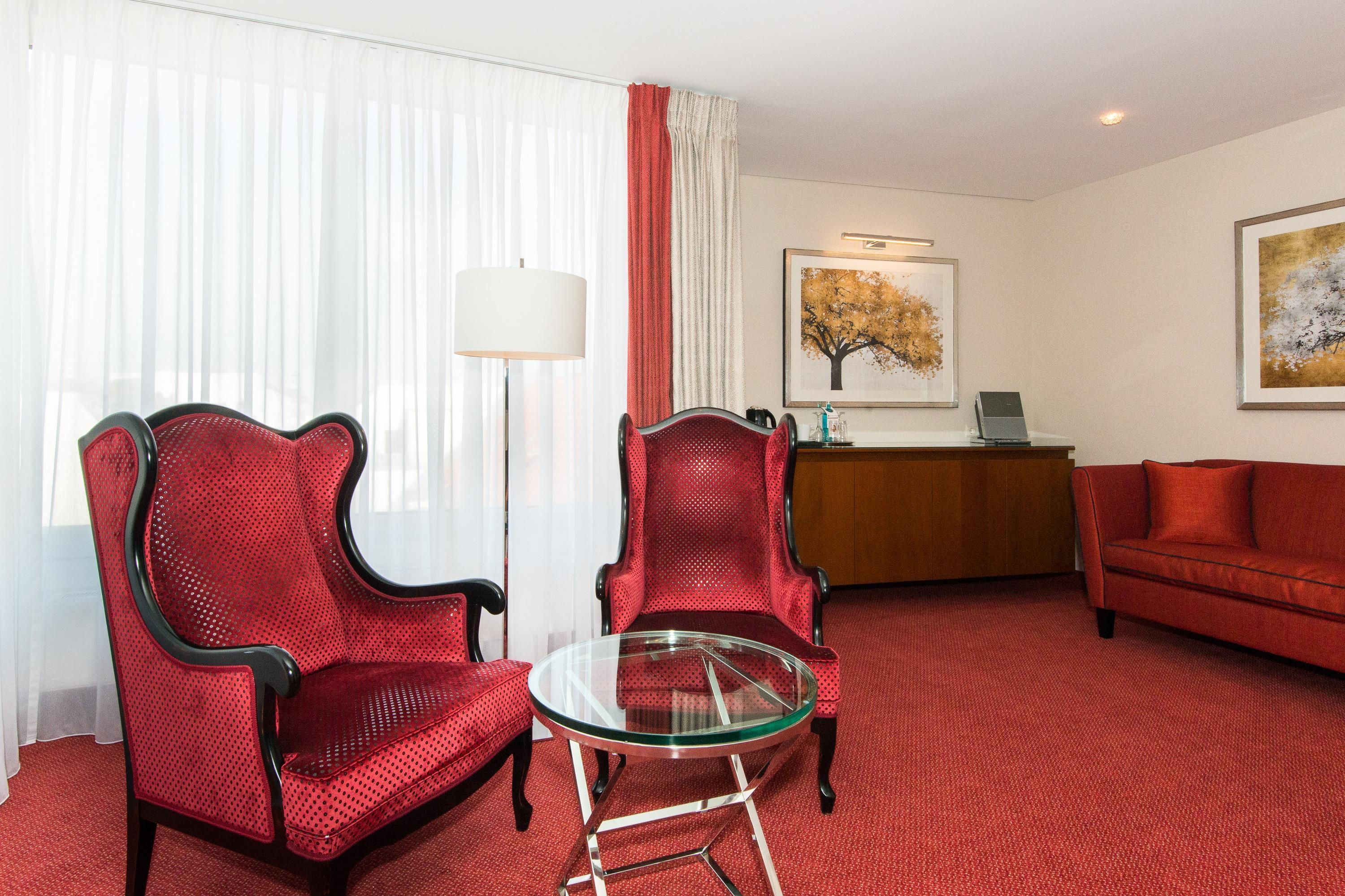 Quarto de hotel com um sofá vermelho, duas poltronas vermelhas, uma mesa de centro de vidro, uma escrivaninha com abajur e um quadro na parede.
