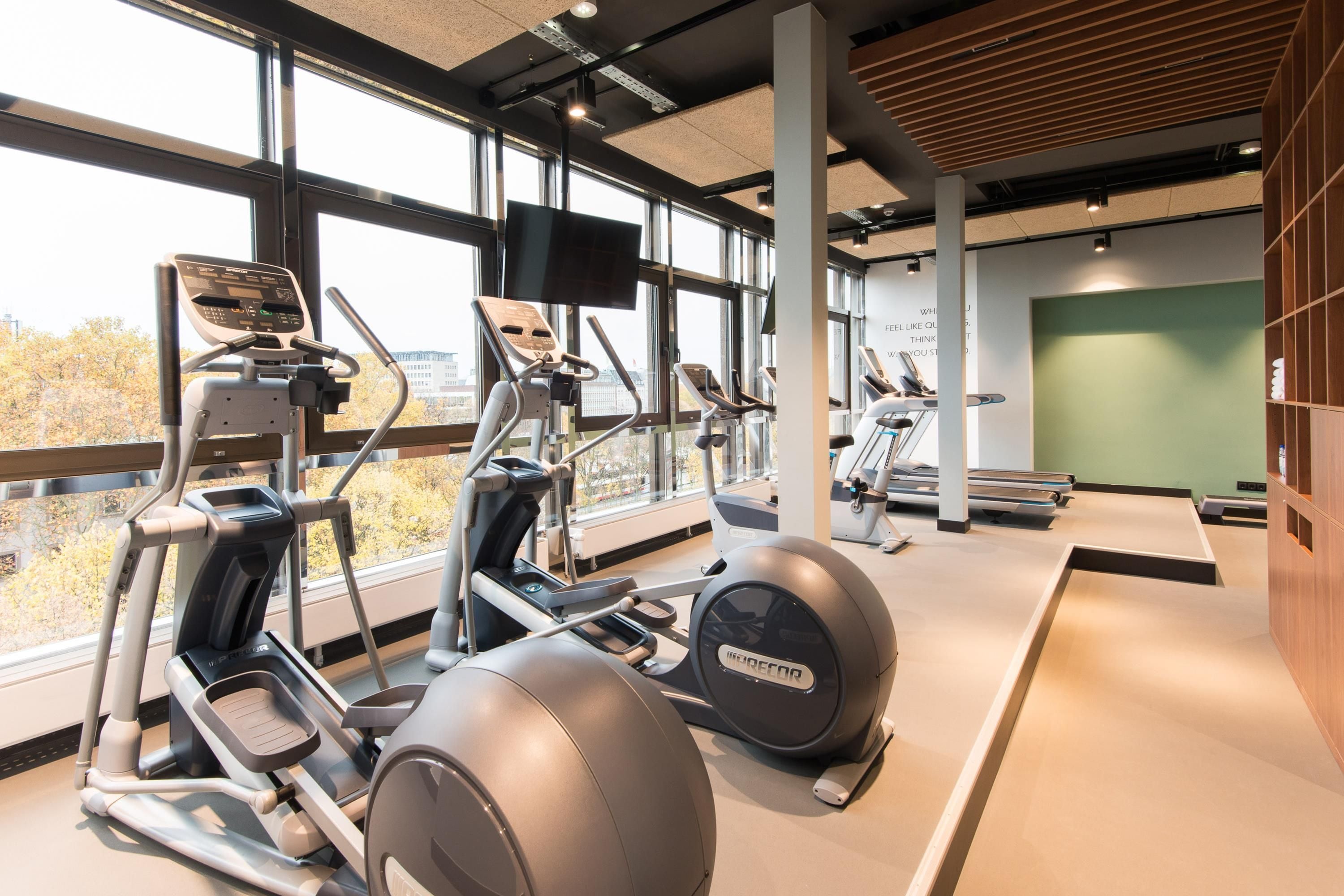 Un gimnasio con equipos de ejercicio variados, ventanas amplias y una pared verde.