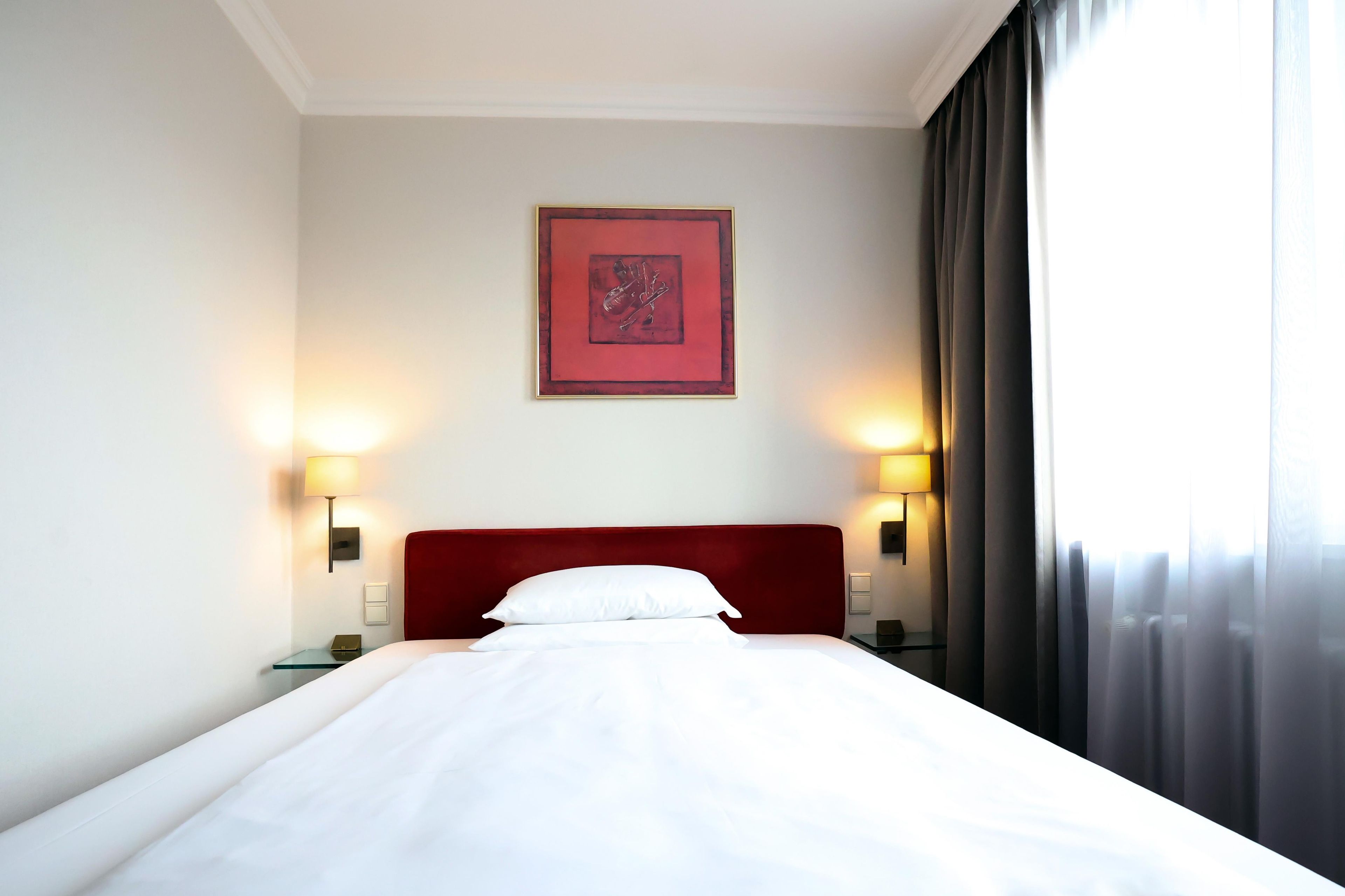 Une chambre d’hôtel avec un lit, deux lampes de chevet, une table de chevet, un tableau encadré au mur et une fenêtre avec des rideaux.