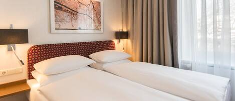 A hotel room with a double bed, two bedside tables, a wall-mounted lamp, a framed picture, and a window with curtains.