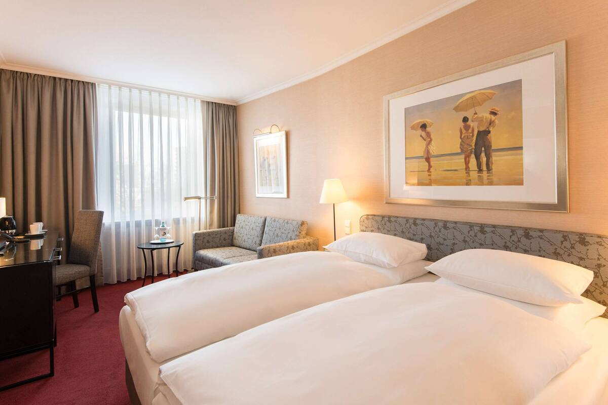 A hotel room with two beds, a desk, a chair, a small table, and a painting on the wall.