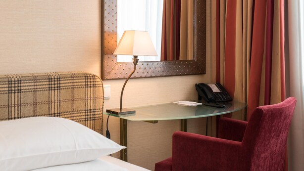 Una habitación de hotel con una cama, un escritorio con una lámpara, un teléfono y una silla roja.