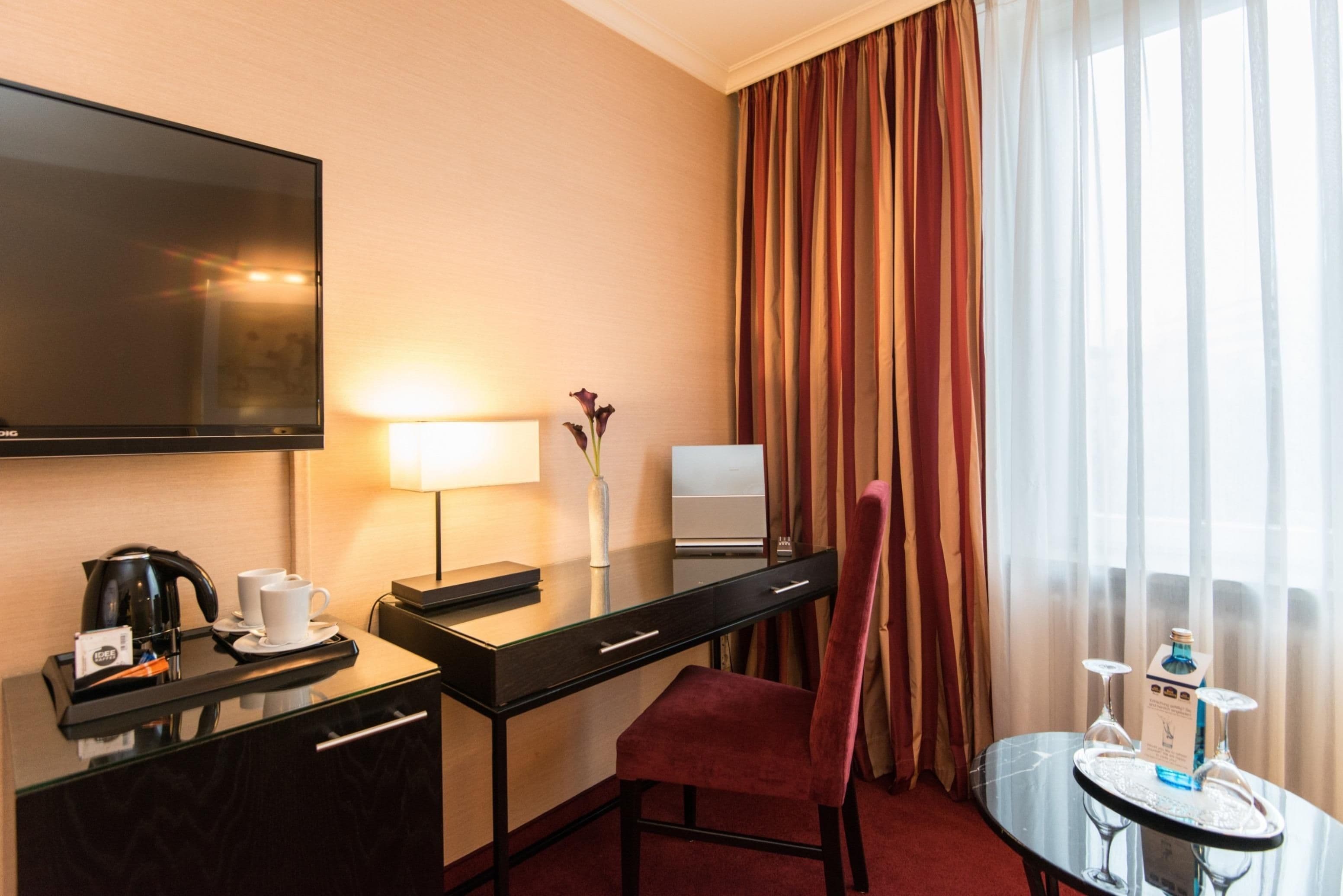 A hotel room with a desk, a chair, a TV, a coffee maker, a lamp, a vase with flowers, a minibar, and a window with curtains.
