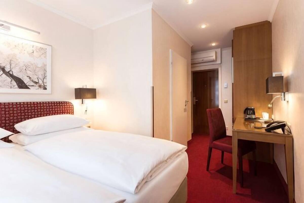 A hotel room with a bed, a desk, a chair, a lamp, and a painting on the wall.