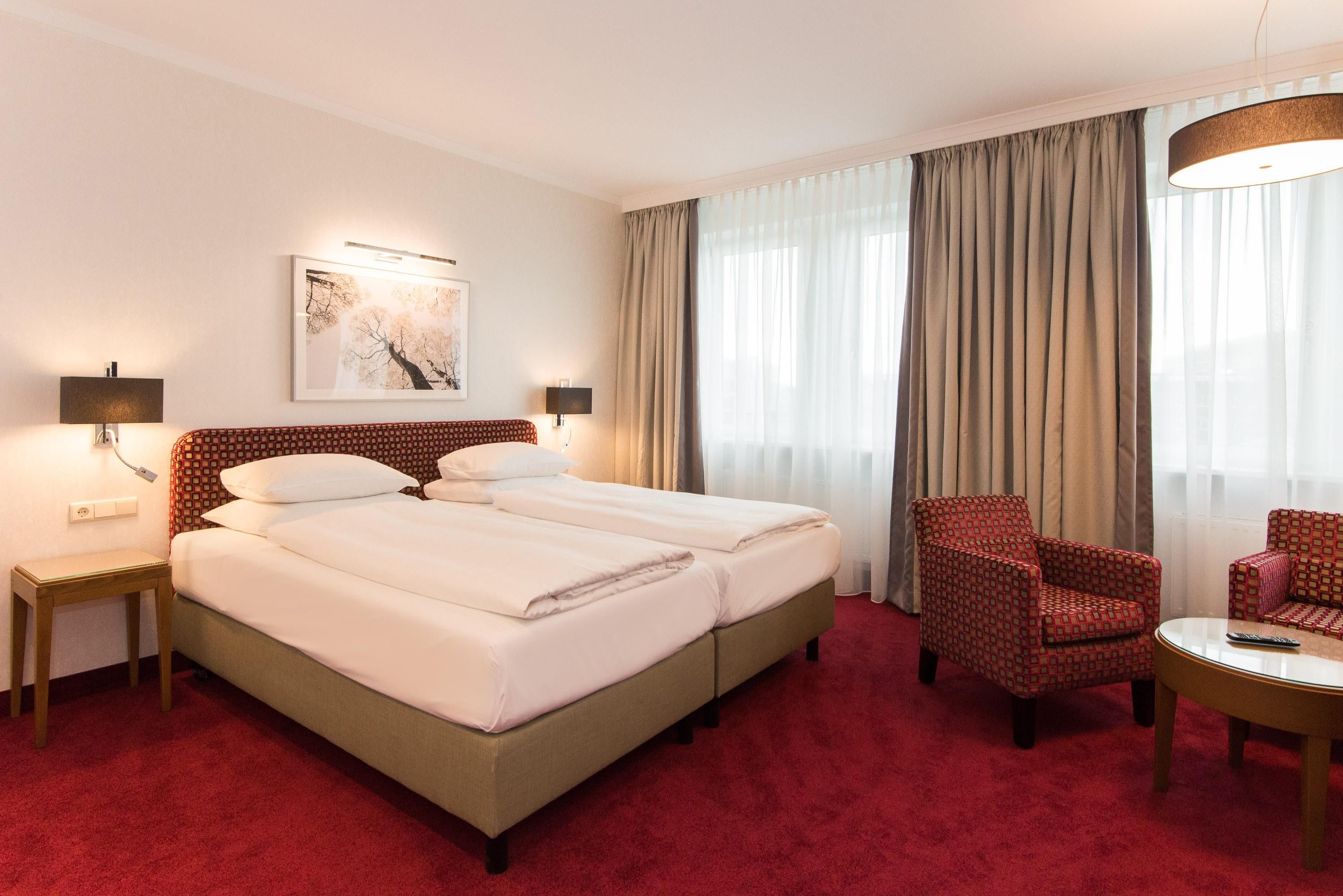 Ein Hotelzimmer mit zwei Betten, einem roten Teppich, einem Sessel, einem kleinen Tisch und einem Bild an der Wand.