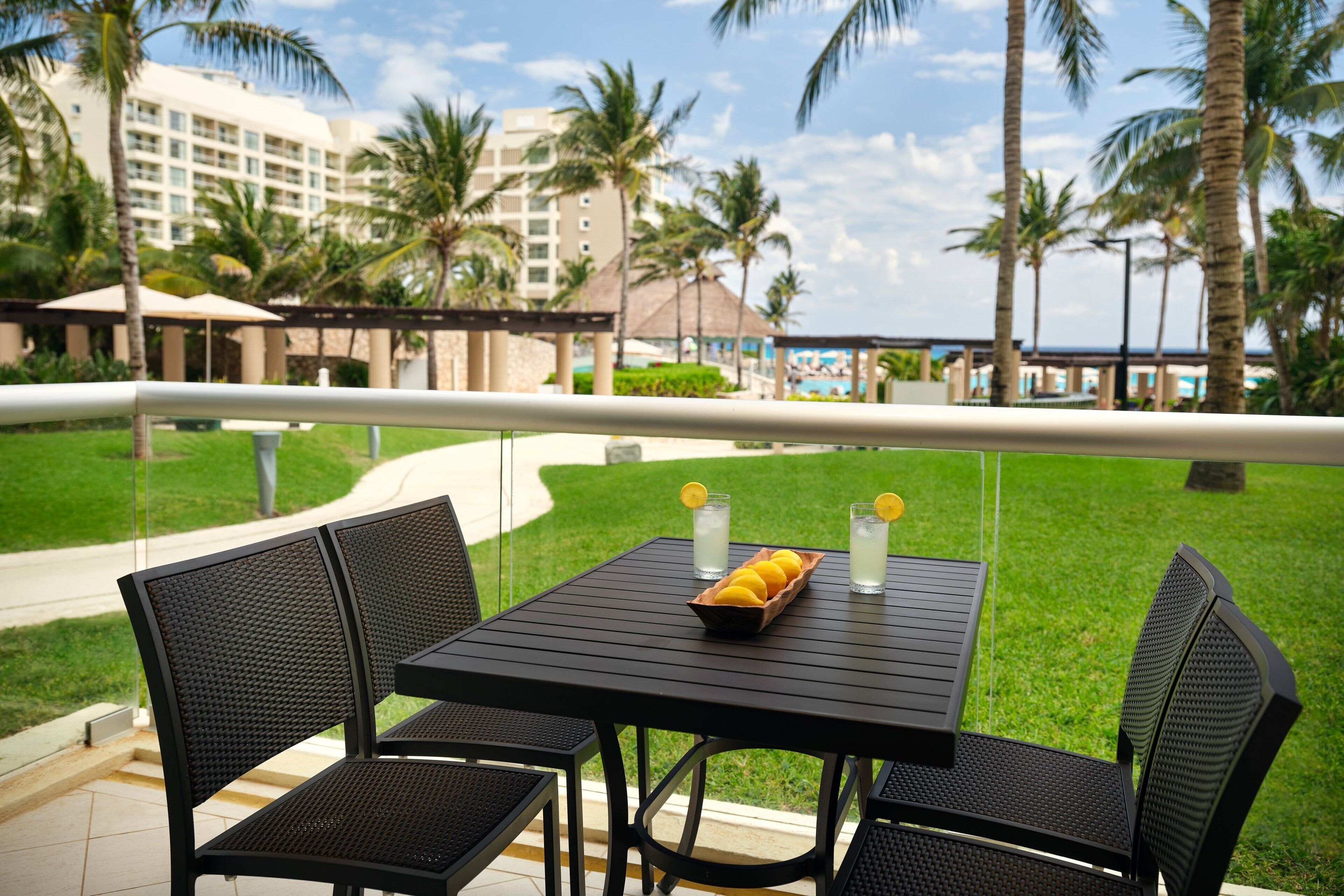 A table with chairs and a tray of lemons and glasses of water on a balcony with a view of palm trees and a resort building.