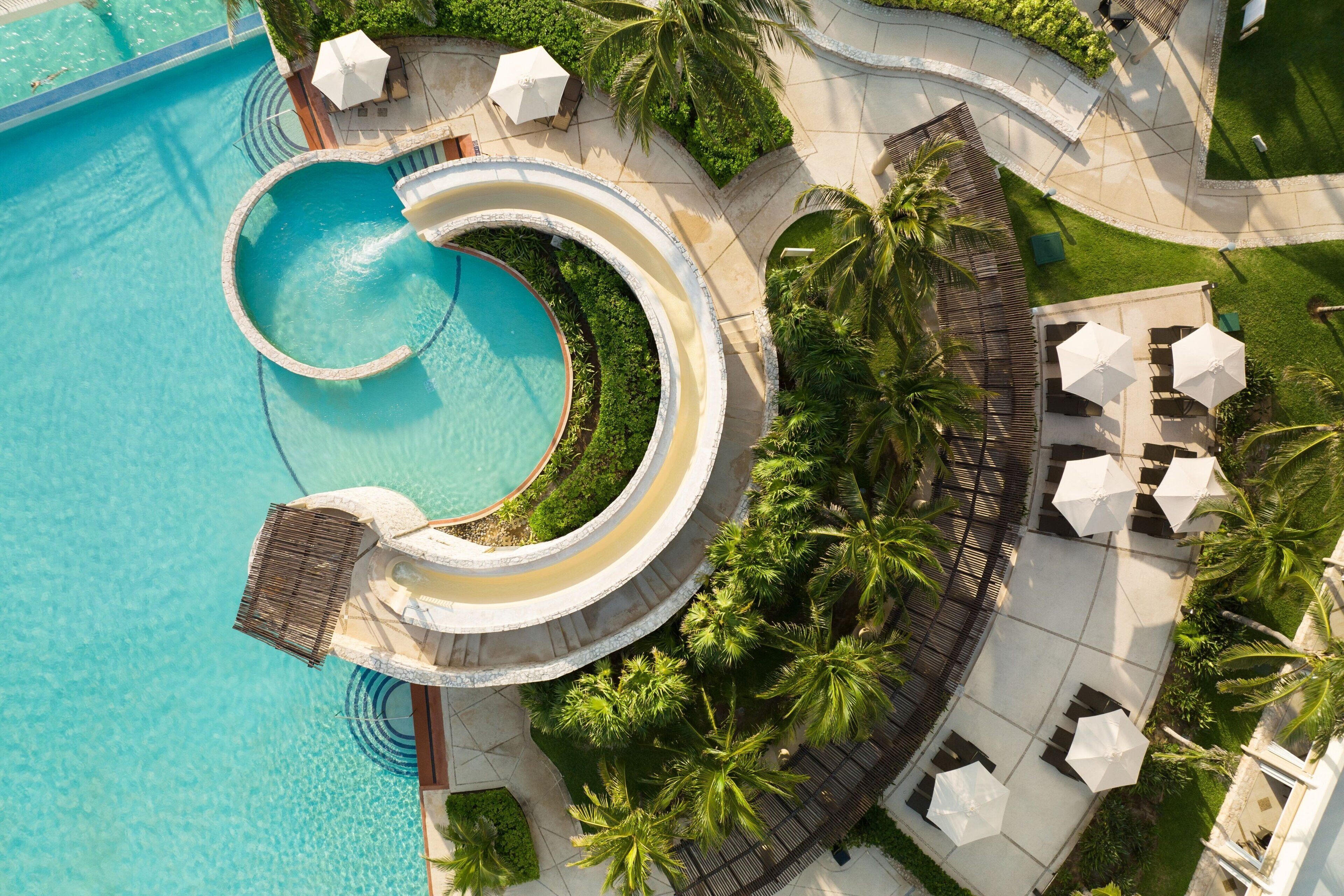 Vue aérienne d’un complexe hôtelier doté d’une piscine circulaire, de palmiers et d’espaces de détente en plein air.