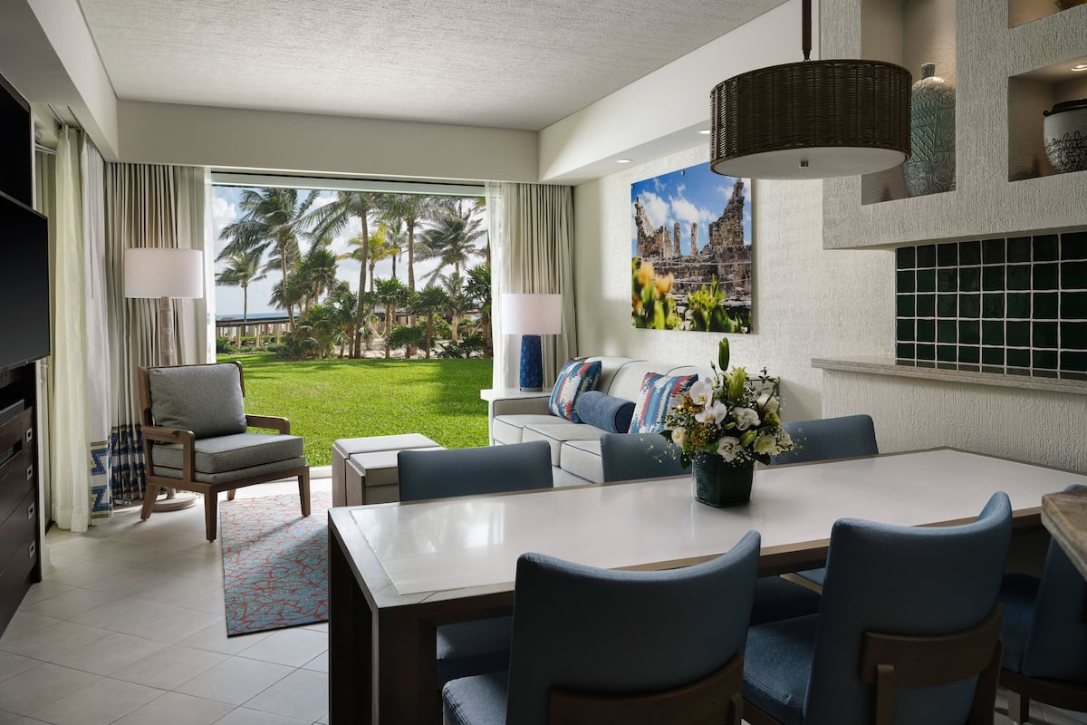 Habitación de hotel moderna con un amplio ventanal que ofrece vistas a un jardín exuberante y palmeras. Cuenta con zona de comedor con mesa y sillas, un conjunto de sofás y un televisor de pantalla plana montado en la pared.
