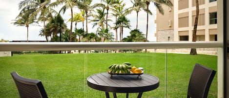 A balcony with two chairs and a table, a bowl of fruit, and a view of palm trees and a building.