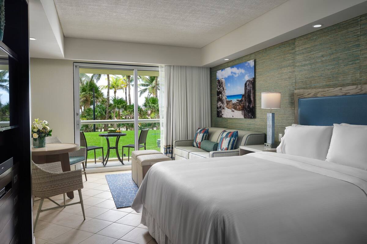 Habitación de hotel con una cama grande, un sofá, una mesa de comedor con sillas, un televisor y vista a una playa y palmeras a través de la puerta de vidrio.