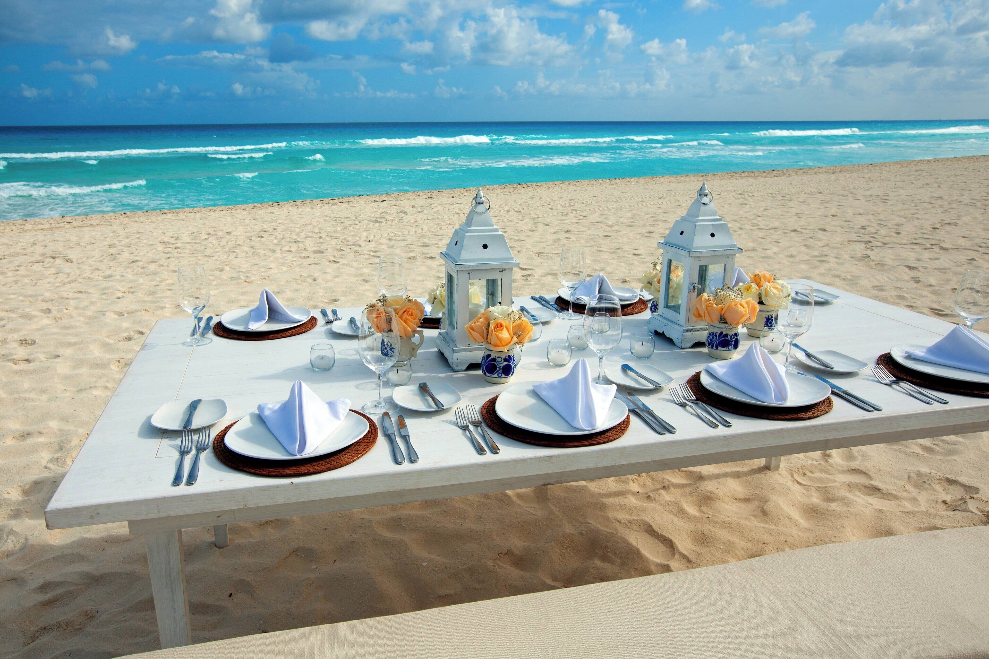 Une table dressée pour deux personnes, avec des nappes blanches, des couverts en argent et des fleurs orange, le tout installé sur une plage avec l’océan en toile de fond et les vagues qui déferlent.