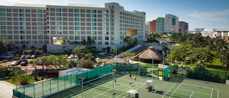 A tennis court surrounded by a hotel complex with multiple buildings, a parking area, and palm trees.