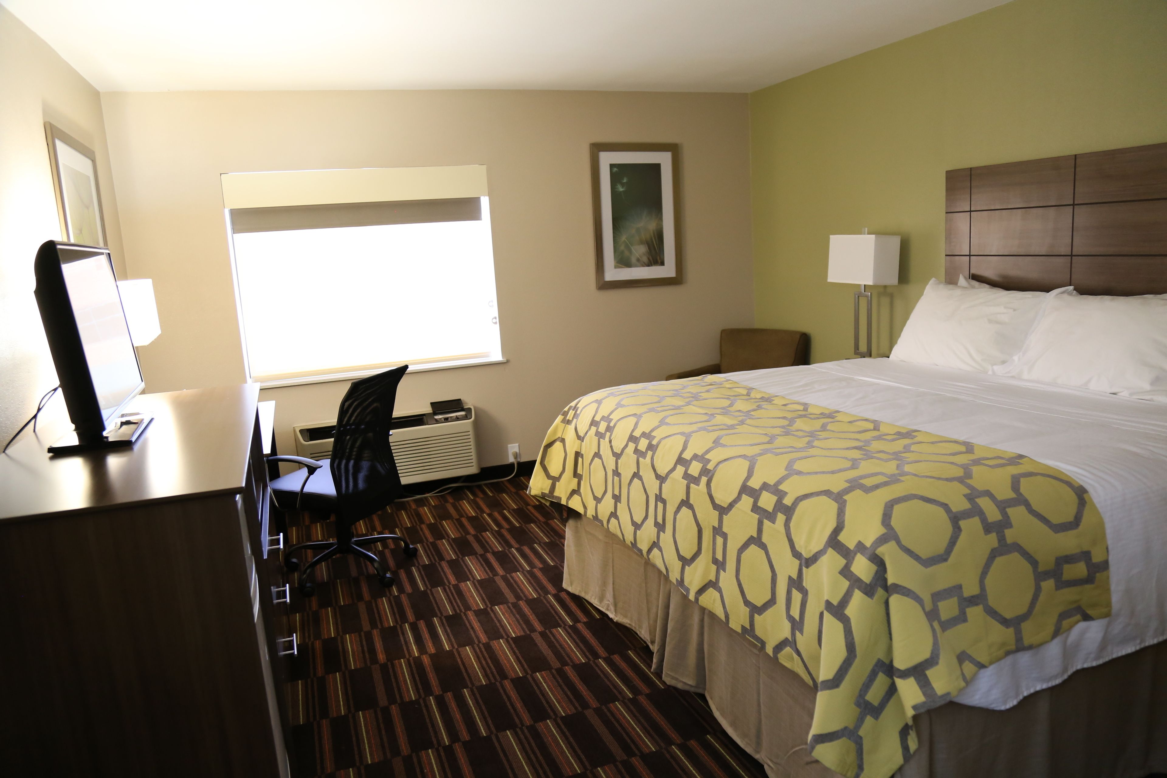 Una habitación de hotel con una cama grande, un escritorio con una computadora, una silla, una ventana y una lámpara de pared.