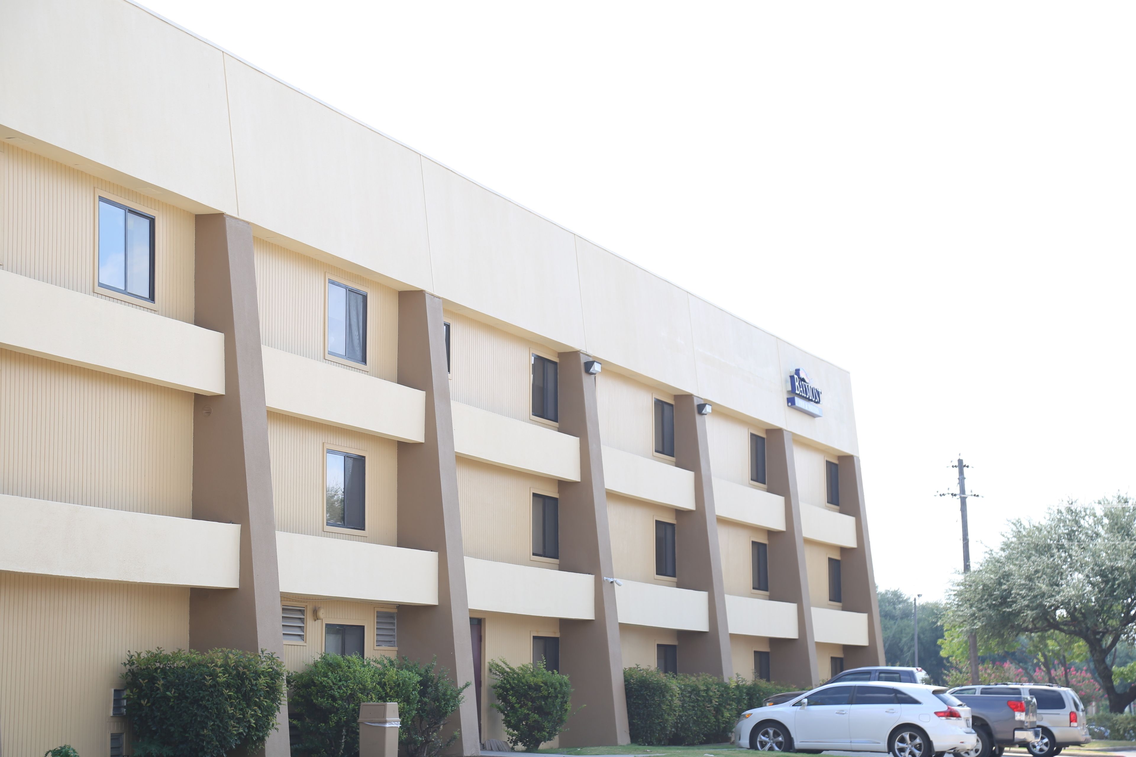 Un hotel de varios pisos con fachada color beige, balcones y un estacionamiento con automóviles.