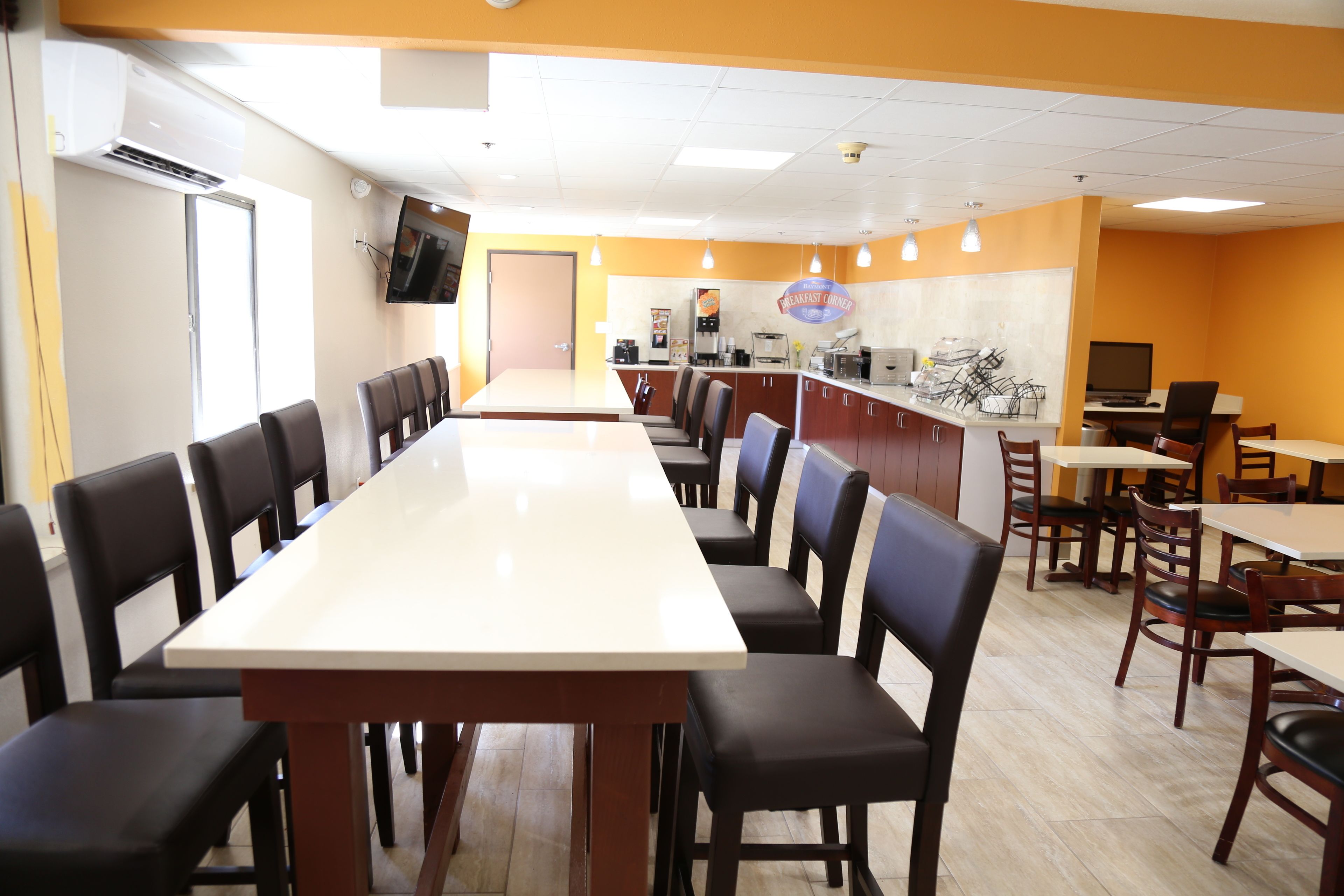 A long dining table with chairs, a counter with a coffee machine, and a TV mounted on the wall.