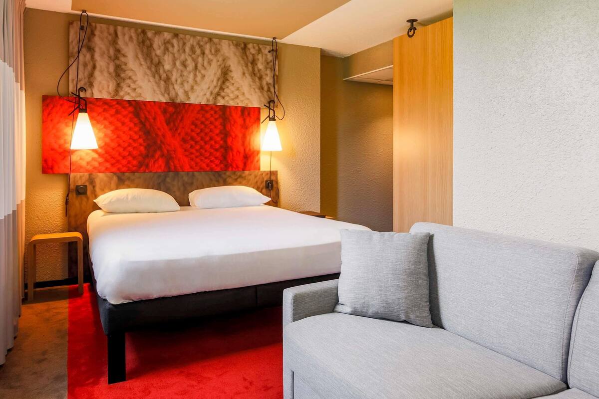 A hotel room with a bed, a grey sofa, a red carpet, and a wall with a red and beige patterned headboard.