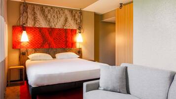 Une chambre d’hôtel avec un lit, un canapé gris, une moquette rouge et un mur orné d’une tête de lit à motifs rouges et beiges.