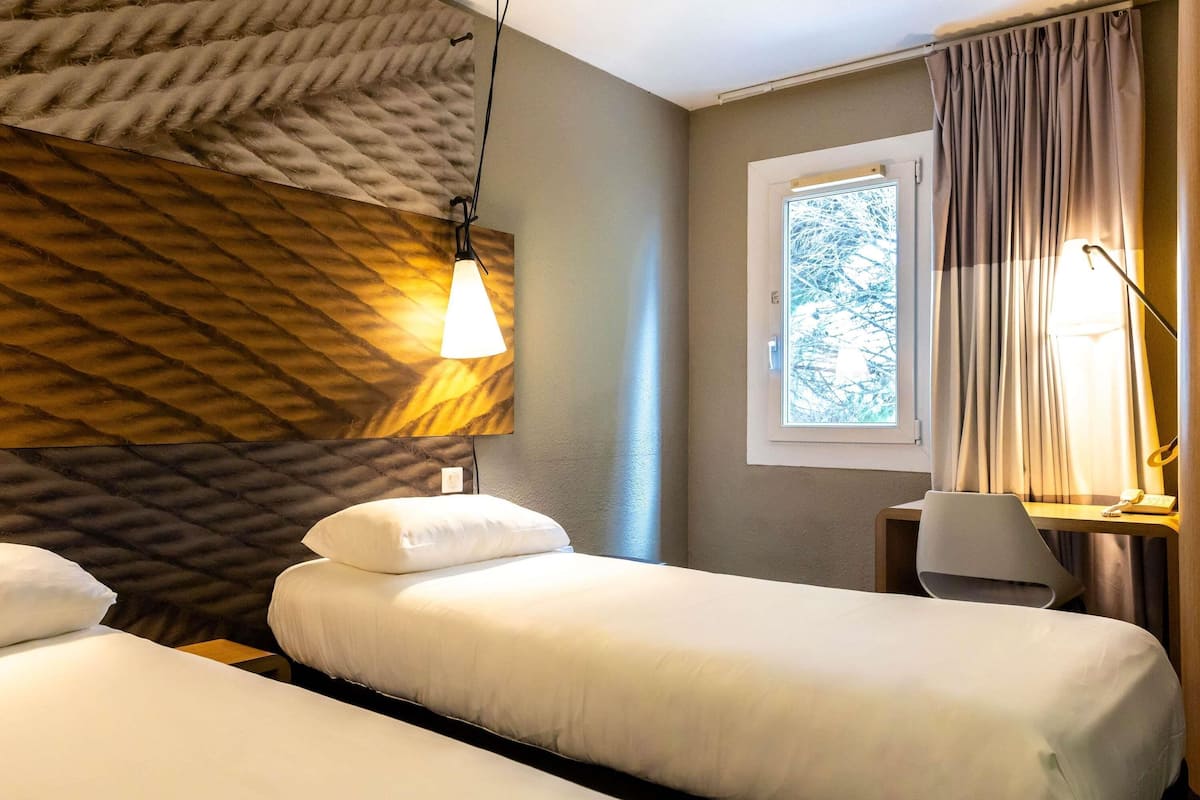 A hotel room with two beds, a window with curtains, a lamp, and a wall with a textured pattern.