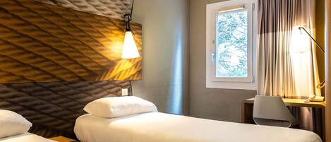 Ein Hotelzimmer mit zwei Betten, einem Fenster mit Vorhängen, einer Lampe und einer Wand mit strukturierter Oberfläche.