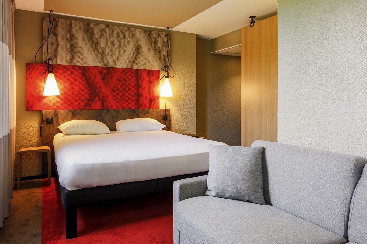 Habitación de hotel con una cama, dos lámparas de noche, alfombra roja, sofá gris y cabecero de madera.
