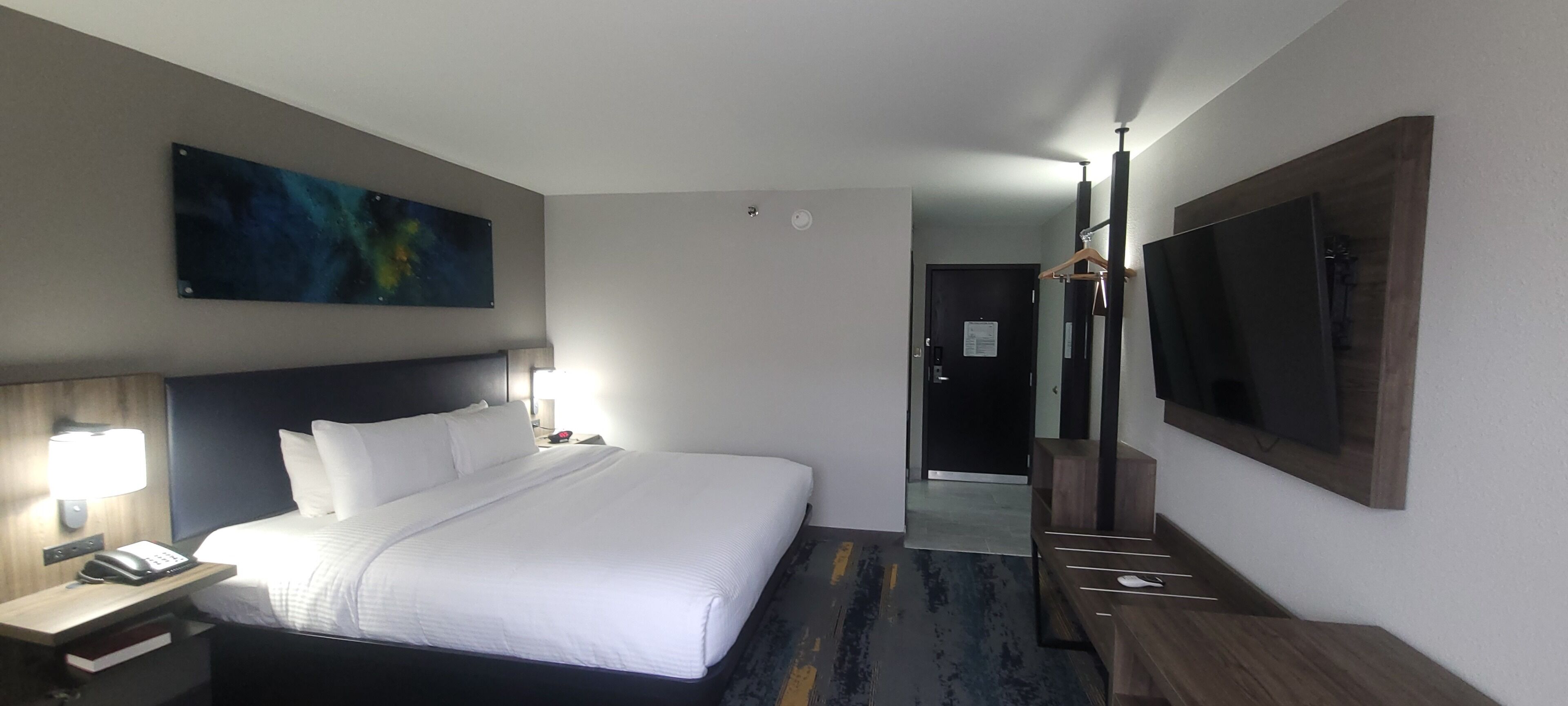 Habitación de hotel con cama, lámparas de noche, escritorio con teléfono, un cuadro grande, espejo y televisor.