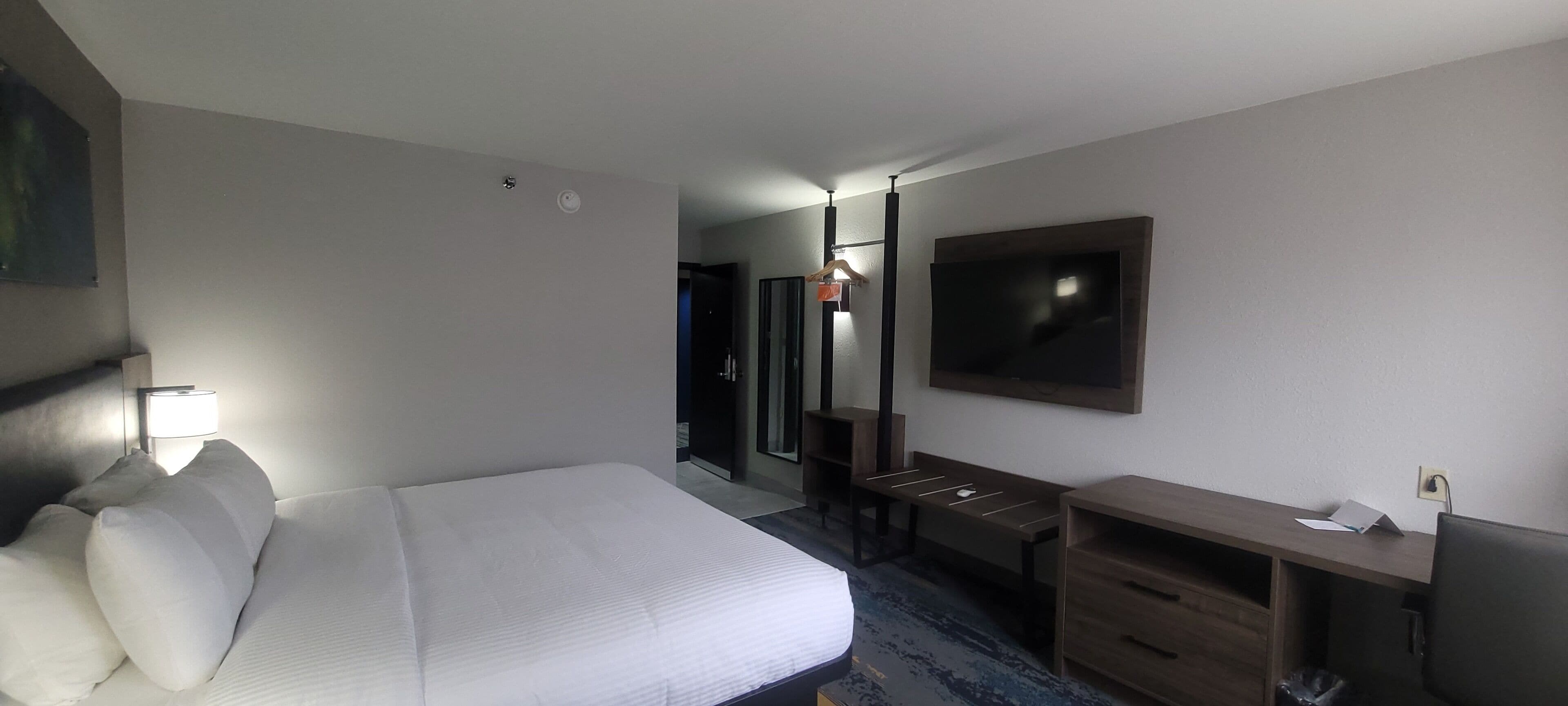 A hotel room with a bed, bedside table, lamp, TV, and a desk with a chair.