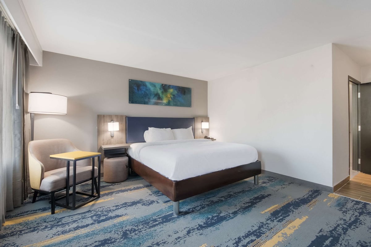 Una habitación de hotel con cama, una silla, una mesita, una mesita de noche y un cuadro en la pared.