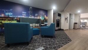 A modern hotel lobby with a cityscape mural, a reception desk, and a seating area with blue chairs and a patterned rug. - Clarion Pointe OKC Airport (Oklahoma City)
