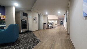 A hotel corridor with a blue chair, a refrigerator, and a trash bin. - Clarion Pointe OKC Airport (Oklahoma City)