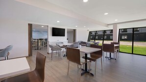 A modern dining area with multiple tables and chairs, a flat-screen TV mounted on the wall, and large windows offering a view of the outside. - Clarion Pointe OKC Airport (Oklahoma City)