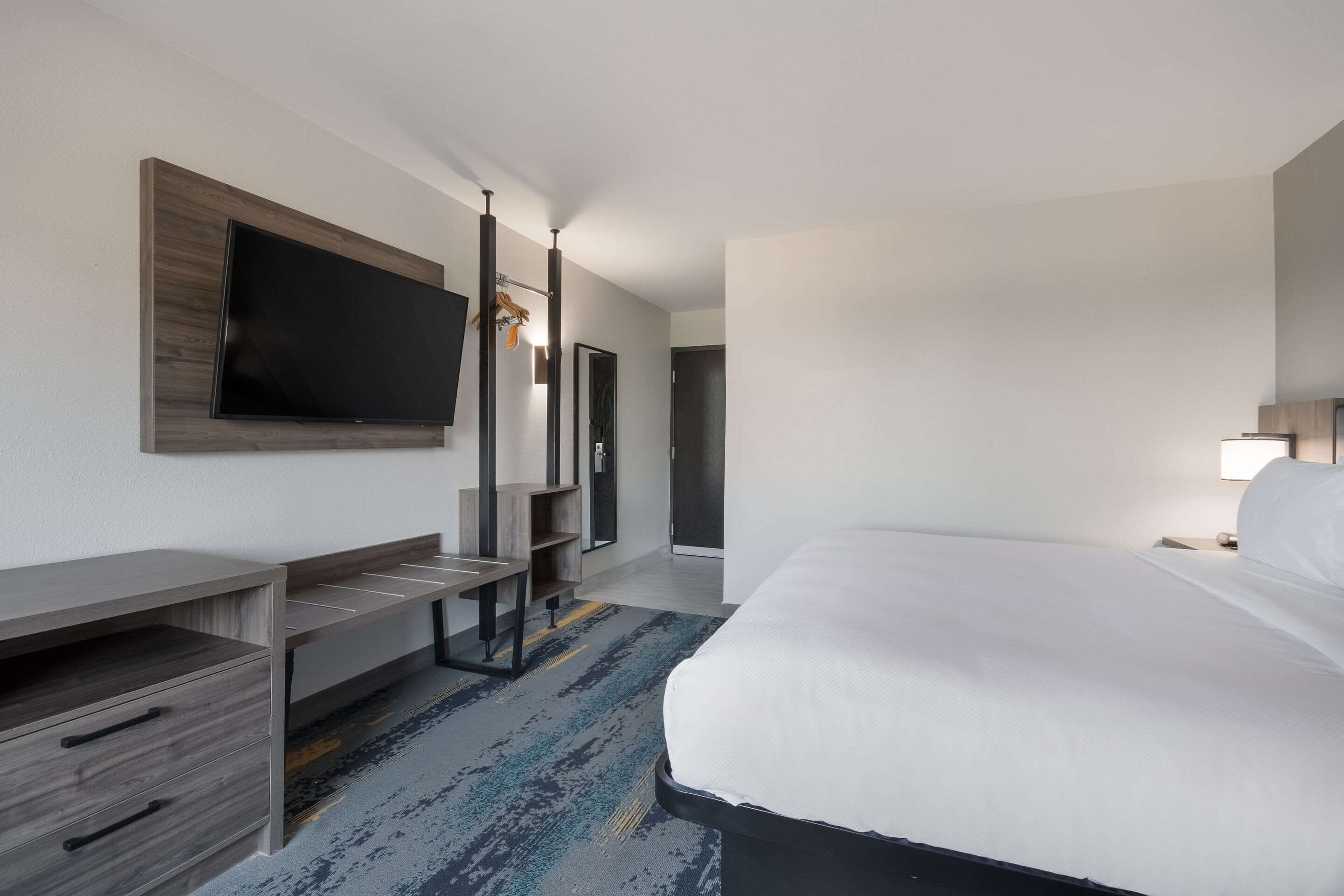 Una habitación de hotel con una cama, un televisor montado en la pared, un escritorio con cajones y un pequeño estante.