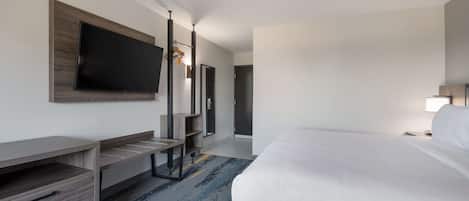 Una habitación de hotel con una cama, un televisor montado en la pared, un escritorio con cajones y un pequeño estante.
