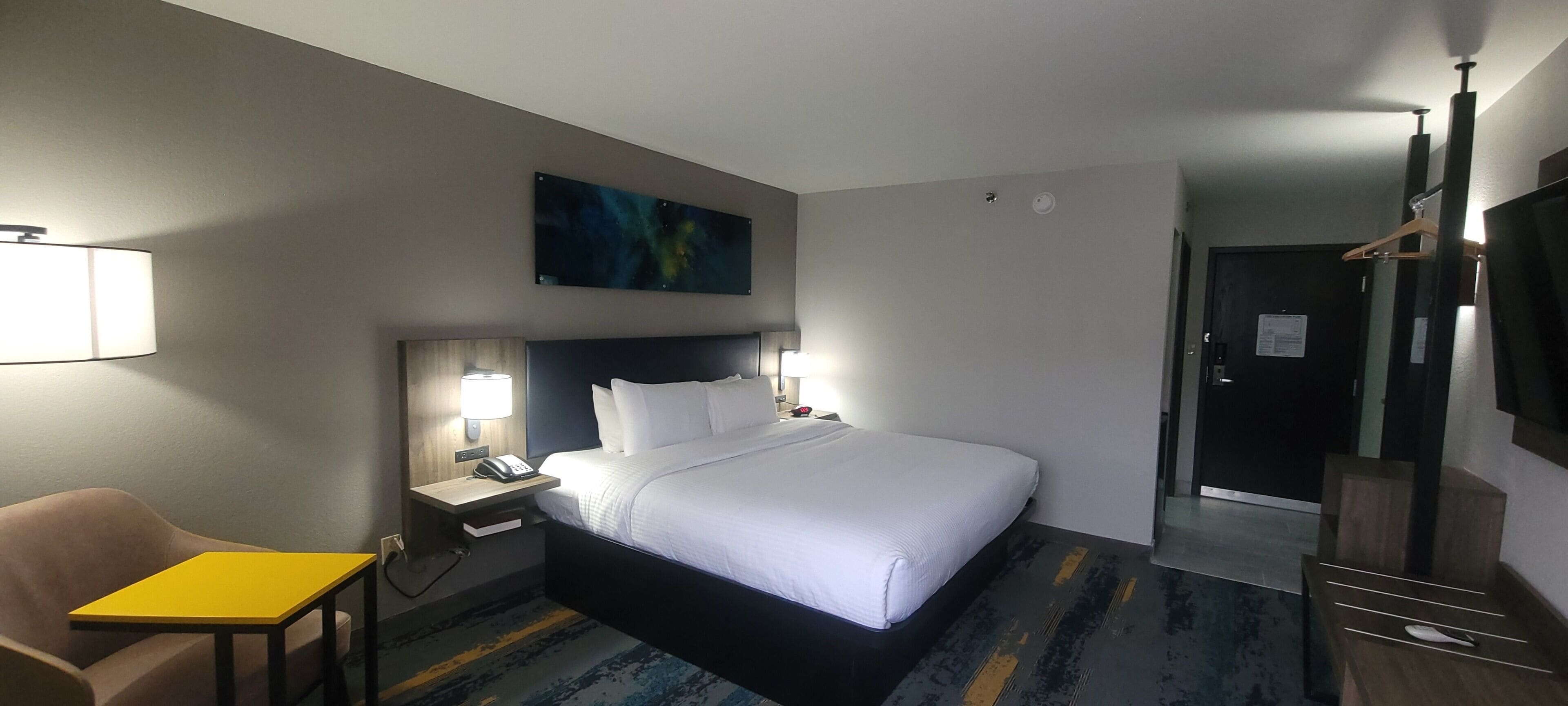 A hotel room with a bed, bedside tables, a chair, a desk, a painting on the wall, and a door.