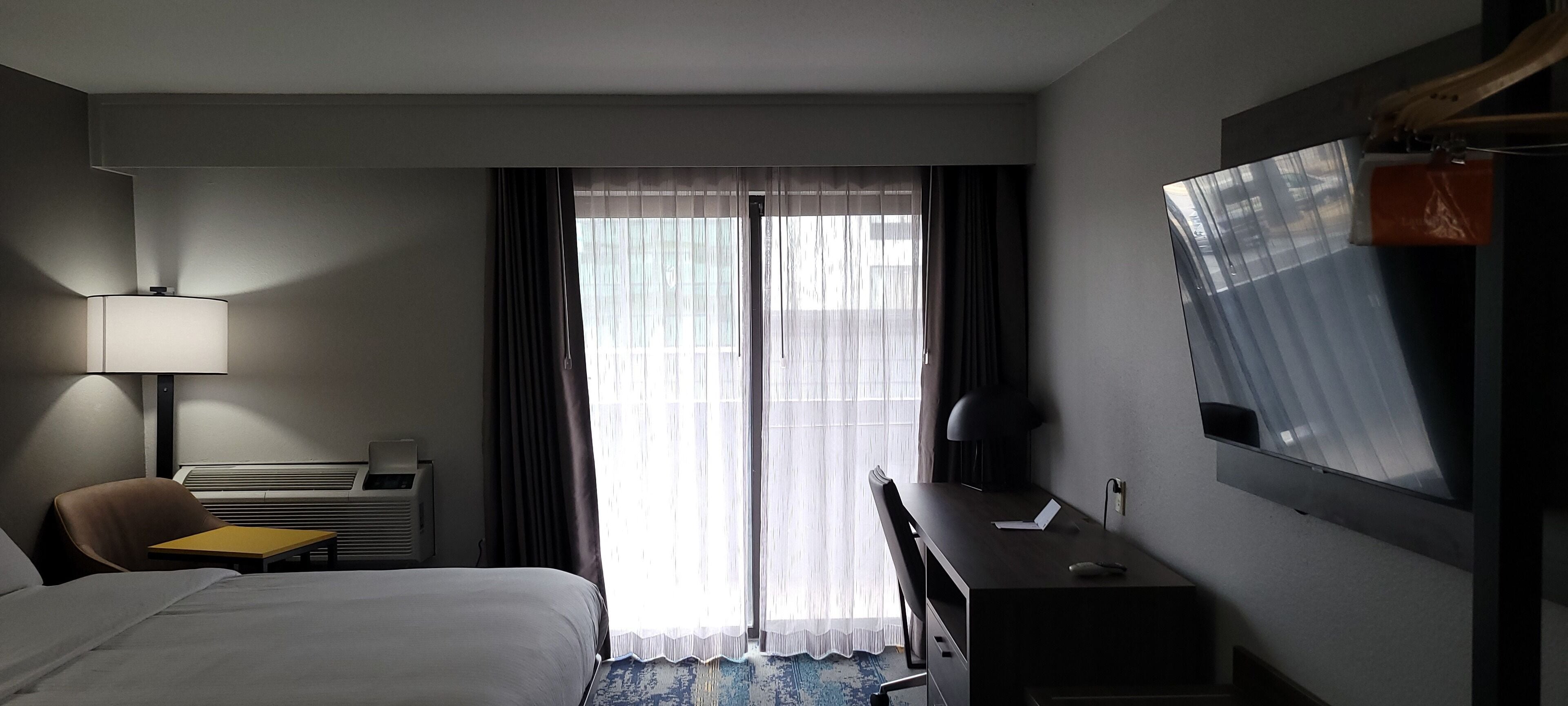 A hotel room with a bed, a desk, a chair, a lamp, a TV, and a window with curtains.