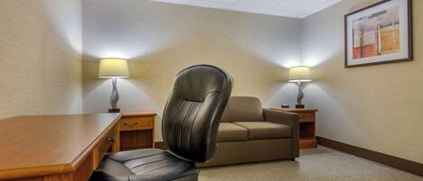 Habitación de hotel con escritorio de madera, sillón de cuero, sofá, dos mesitas de noche con lámparas y un cuadro en la pared.