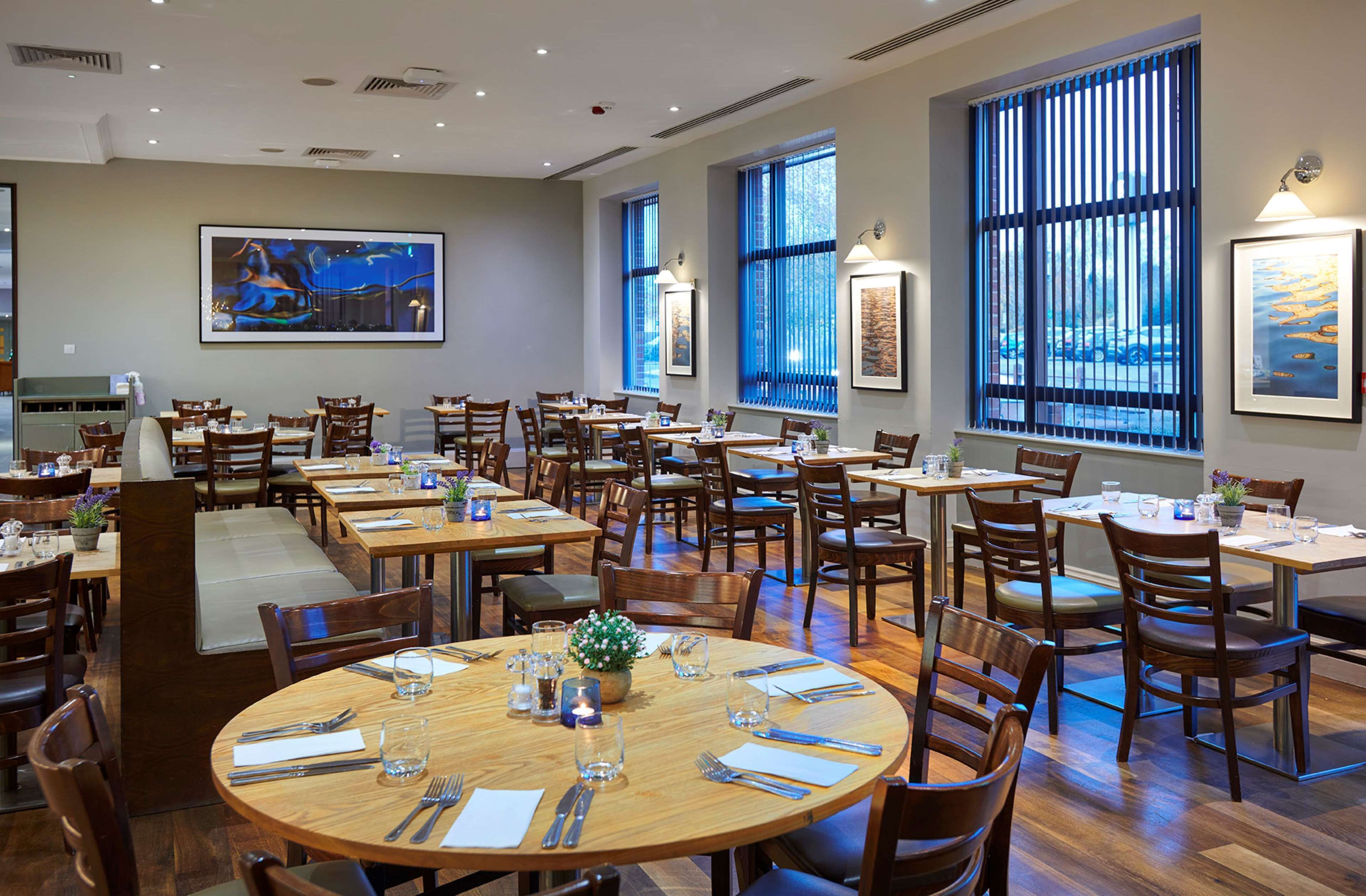 A spacious dining area with multiple tables and chairs, large windows with blinds, and artwork on the walls.