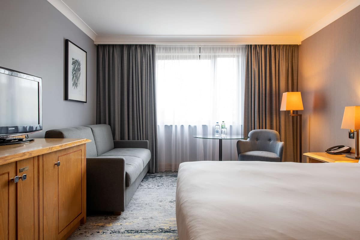 A hotel room with a bed, a sofa, a desk with a lamp, a chair, and a TV.