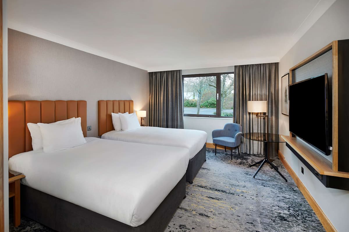 A hotel room with two beds, a TV, a chair, and a window with curtains.