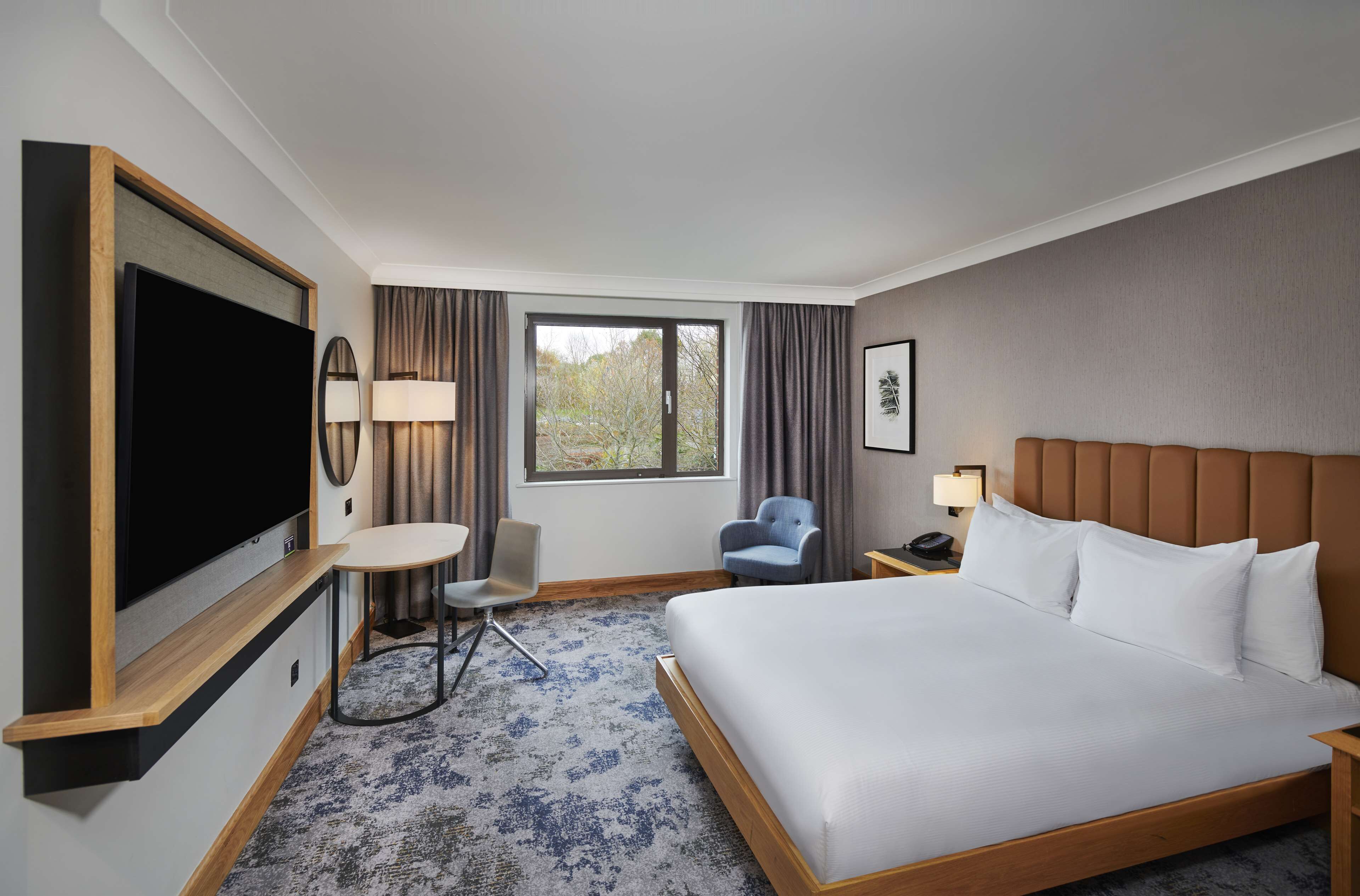 Een hotelkamer met een groot bed, een flatscreen televisie bevestigd aan een houten console, een klein rond tafeltje met een stoel, een bureau met een lamp en een raam met gordijnen.