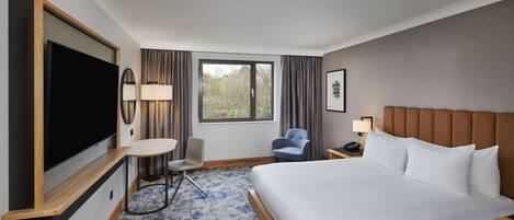 Een hotelkamer met een groot bed, een flatscreen televisie bevestigd aan een houten console, een klein rond tafeltje met een stoel, een bureau met een lamp en een raam met gordijnen.