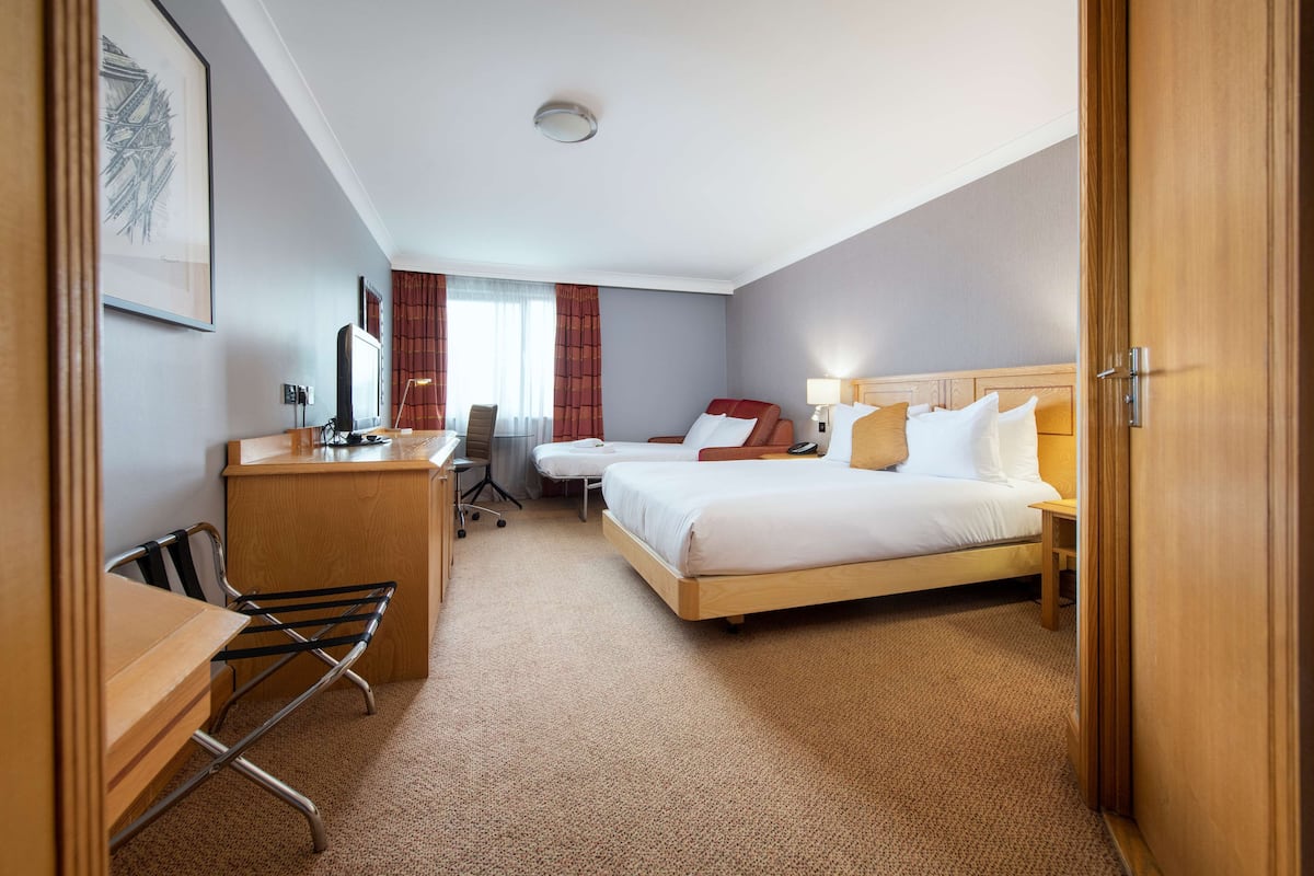 A hotel room with a desk, two beds, a chair, and a window with curtains.
