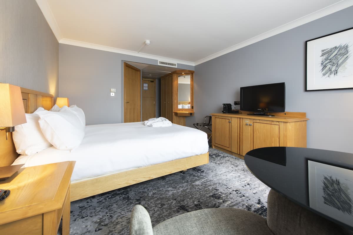 A hotel room with a bed, bedside table, TV, wooden wardrobe, and a desk with a chair.