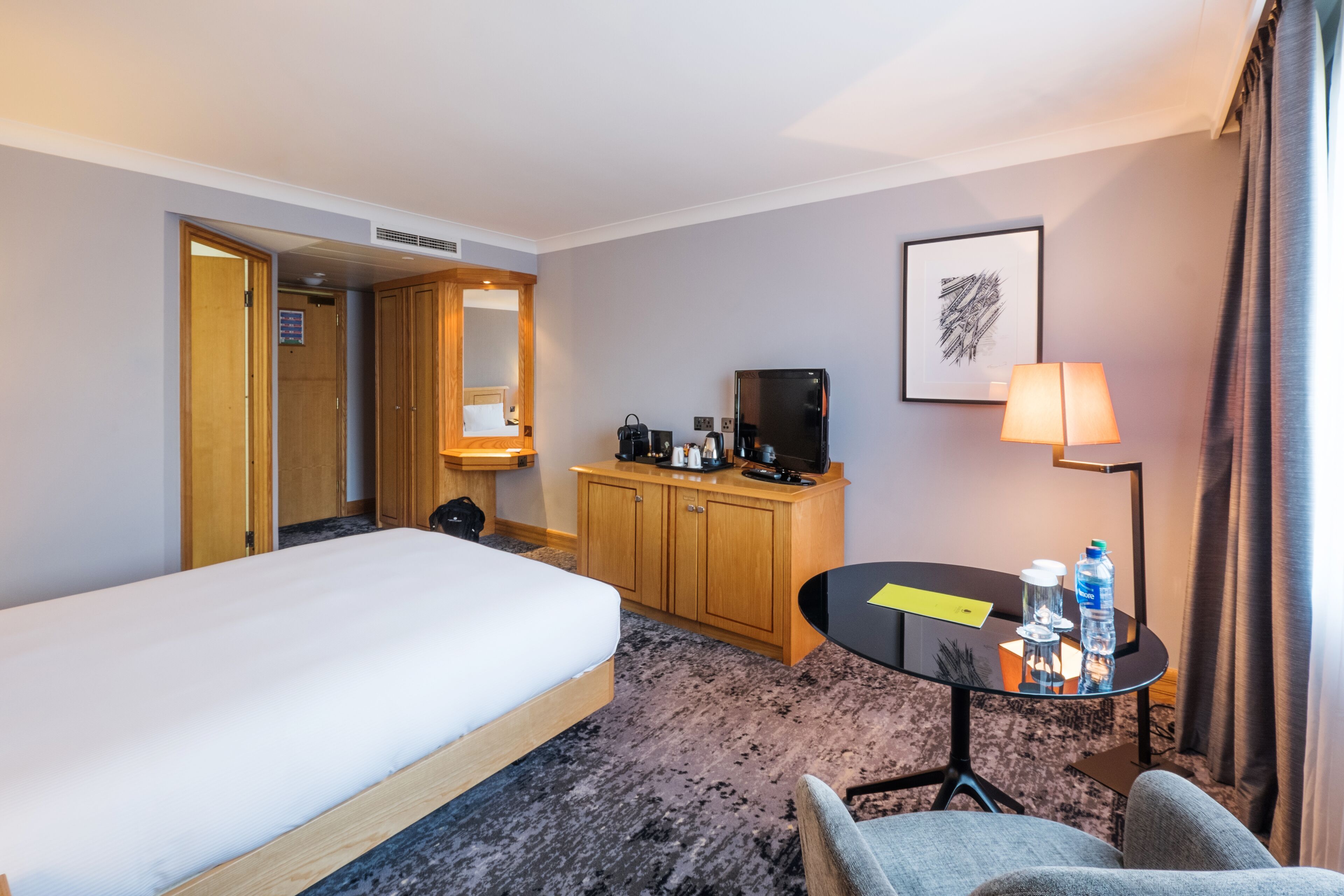A hotel room with a bed, a desk with a lamp, a television, and a small table with water bottles.
