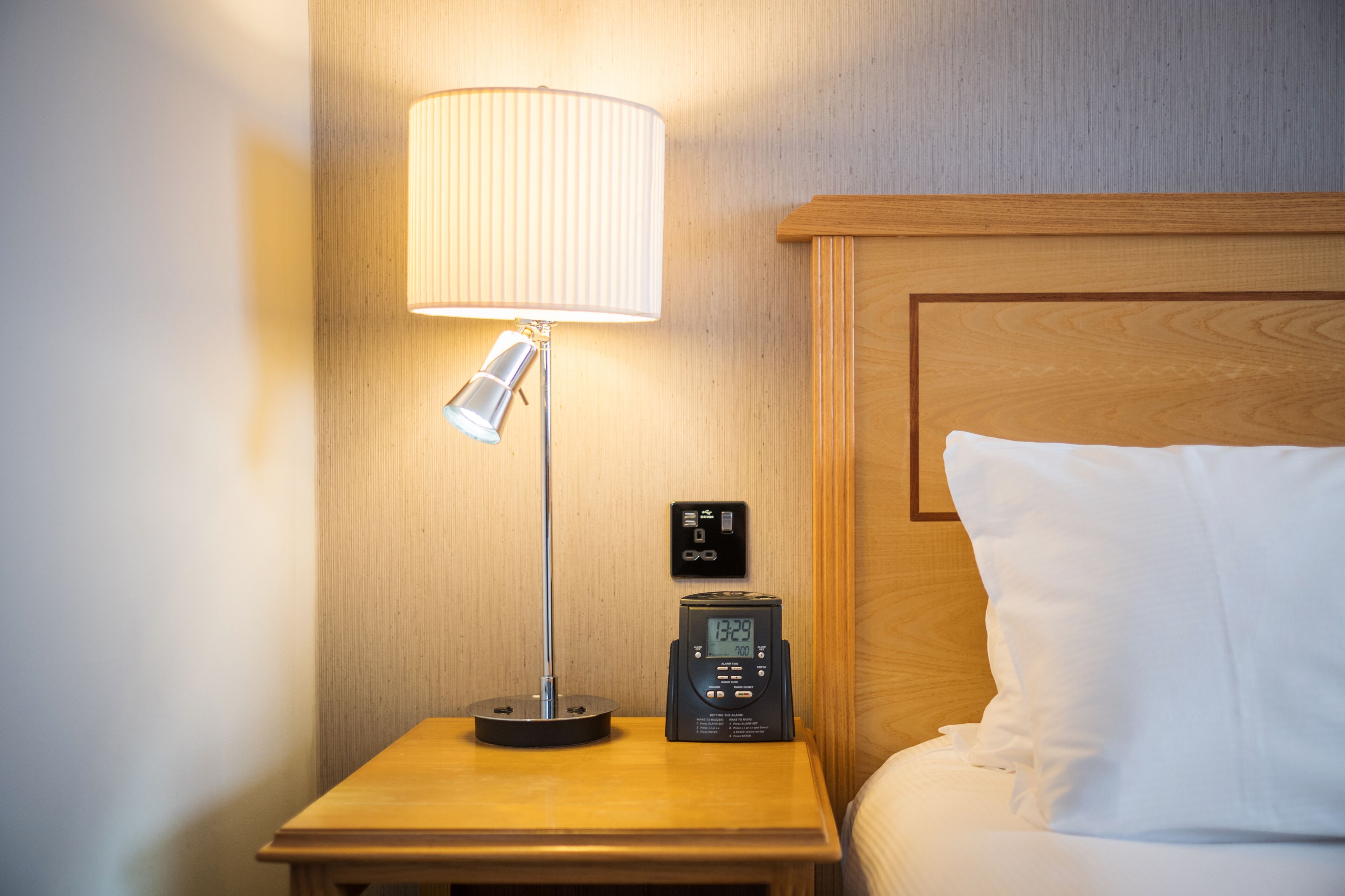 A wooden bedside table with a lamp, a digital clock, and a power outlet.