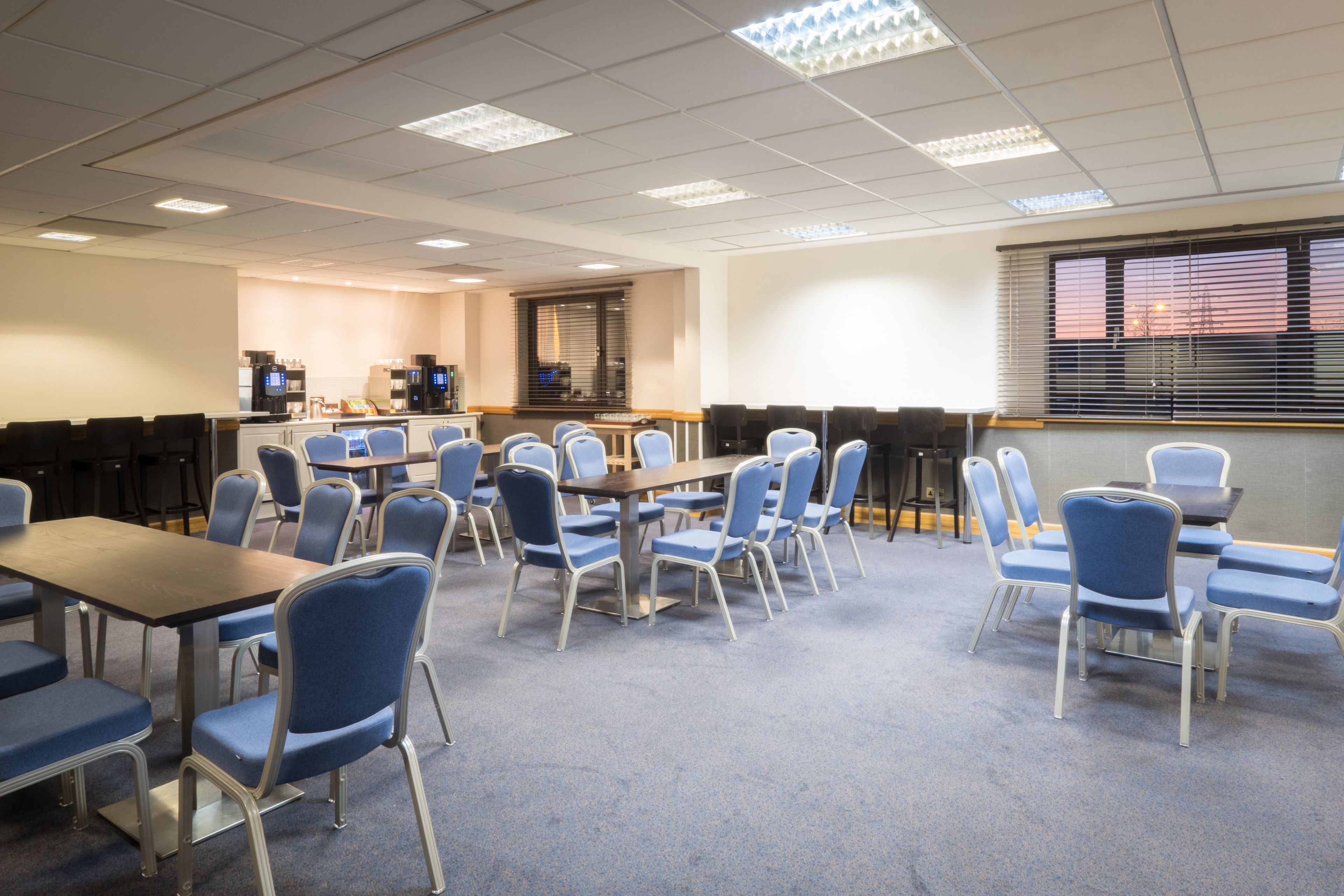 A conference room with multiple tables and chairs, a kitchenette area, and a large window with blinds.