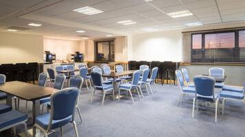 A conference room with multiple tables and chairs, a kitchenette area, and a large window with blinds.