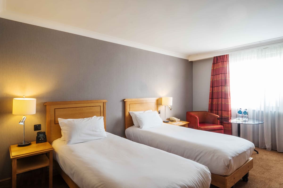 A hotel room with two beds, a nightstand with a lamp, a chair, a small table with a phone and water bottles, and a window with curtains.