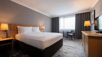 A hotel room with a large bed, two bedside lamps, a desk with a chair, a television, and a window with curtains.