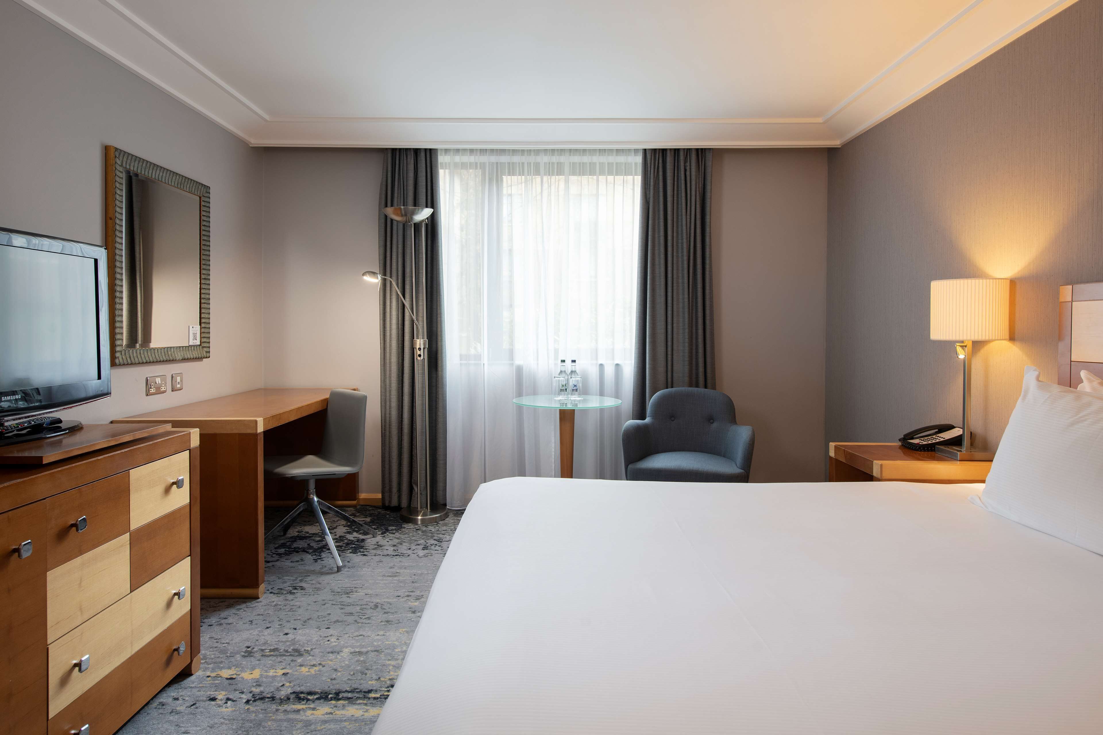 Una habitación de hotel con cama, escritorio, silla, televisor, lámpara y teléfono en la mesita de noche.